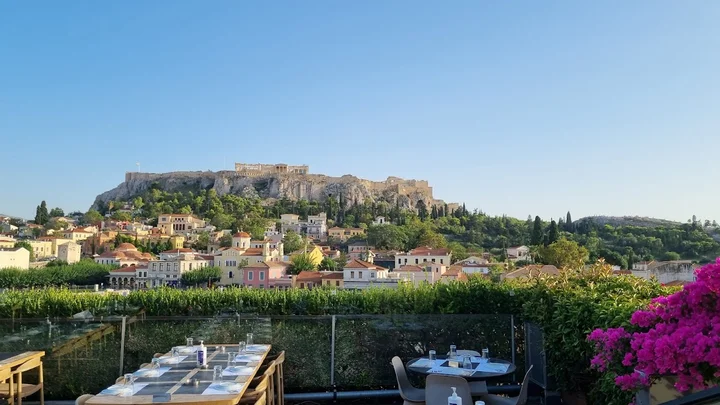 View of the Acropolis of Athens from a rooftop, with historic Greek buildings, a tree-lined hillside and colourful bougainvillea in the foreground