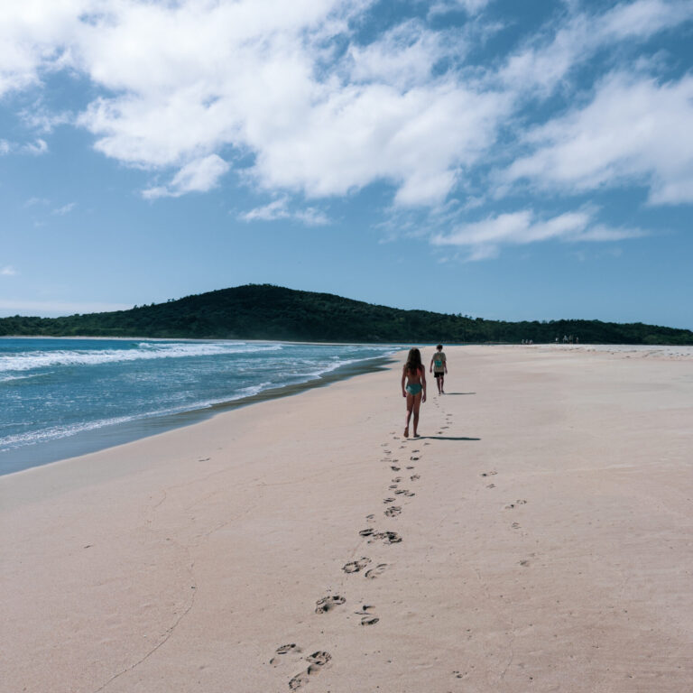 Two kids walking along a quiet sandy beach in Port Stephens, NSW, leaving footprints in the sand with ocean waves and costal headland in the background.