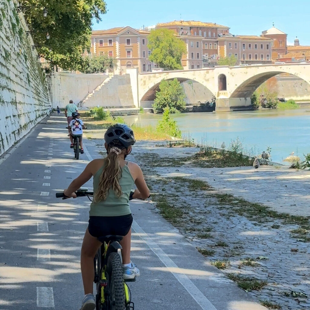 Family cycling along the Tiber River in Rome, Italy, past the historic Ponte Sisto stone bridge and riverside walking and cycling paths