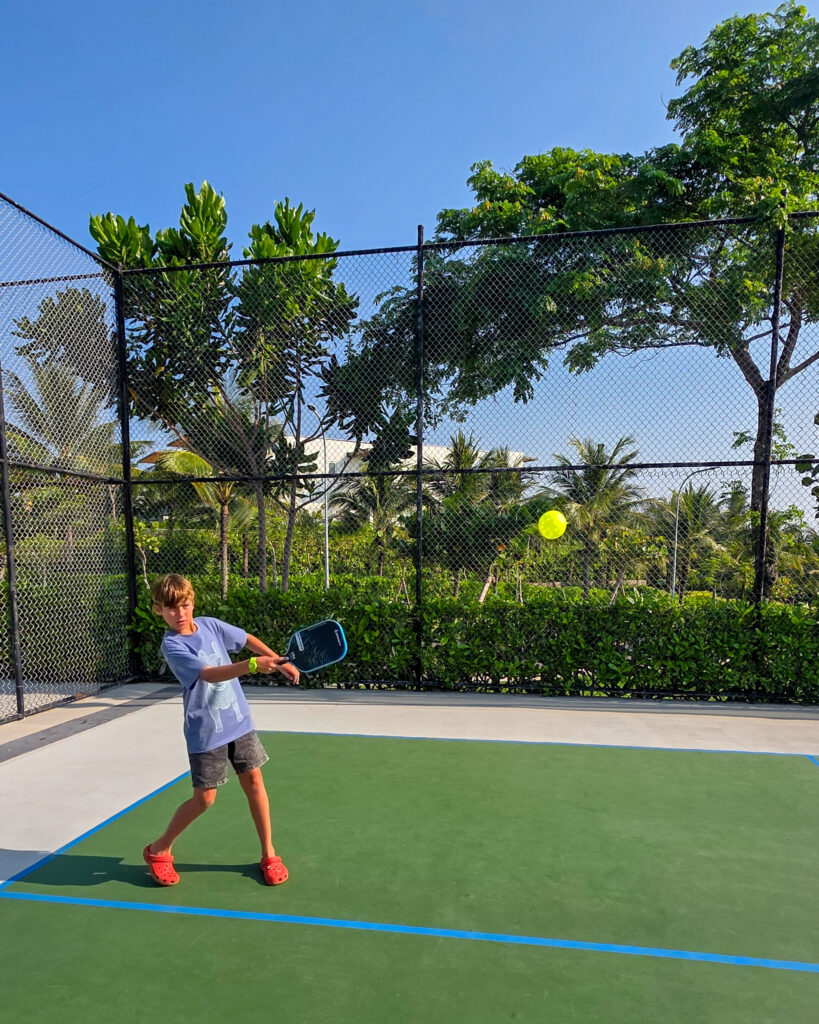 Pickleball court at Alma Resort in Vietnam, with a child playing pickleball surrounded by tropical gardens and palm trees on a family holiday
