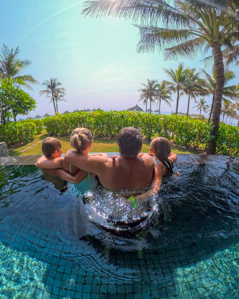 Family relaxing together in the infinity pool at Alma Resort, Vietnam, overlooking the palm-lined beach and ocean on a family holiday